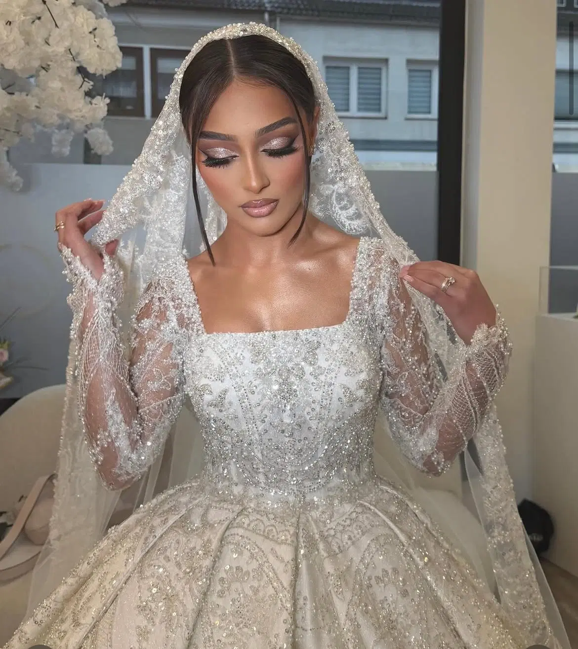Mariée en robe princesse blanche entièrement brodée de perles et de sequins, à décolleté carré et manches longues transparentes, sous un voile perlé