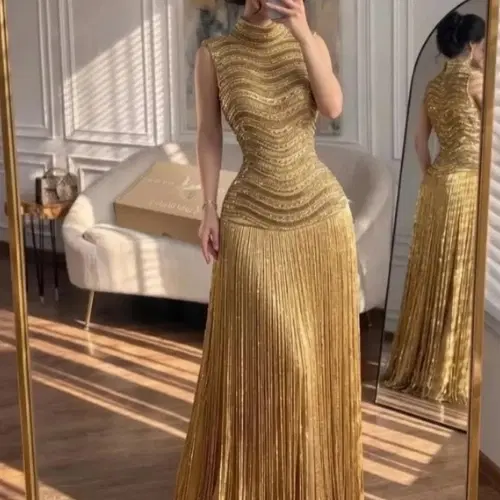 Femme en longue robe de soirée dorée à col montant, buste brodé de perles et jupe à franges, devant un miroir