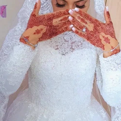 Mariée en robe blanche de dentelle perlée et voile brodé, les mains ornées de henné croisées devant le visage